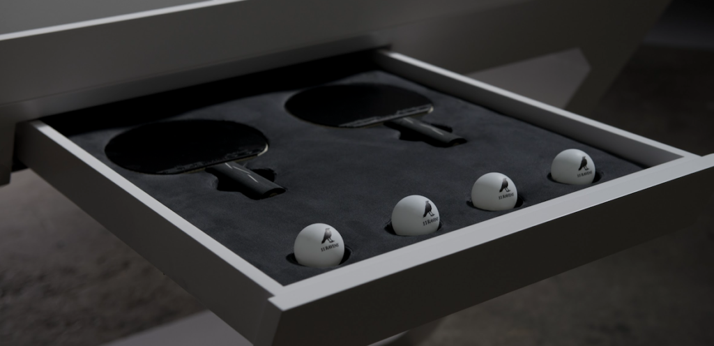 Beneath the Avettore Lucite Ping Pong Table, a drawer holds two black paddles and four white balls, all featuring the SPONETA logo. The balls are neatly arranged in the soft interior, adding elegance to your gaming experience.