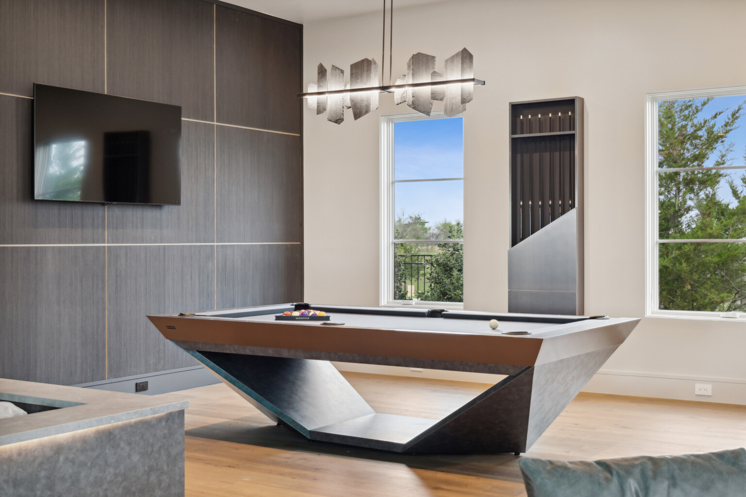 A modern game room exudes elegance with the Stealth Pool Table at its center. It features luxury game tables, a wall-mounted TV, a cue rack, and large windows with tree views, while a contemporary chandelier hangs above the lavish setup.