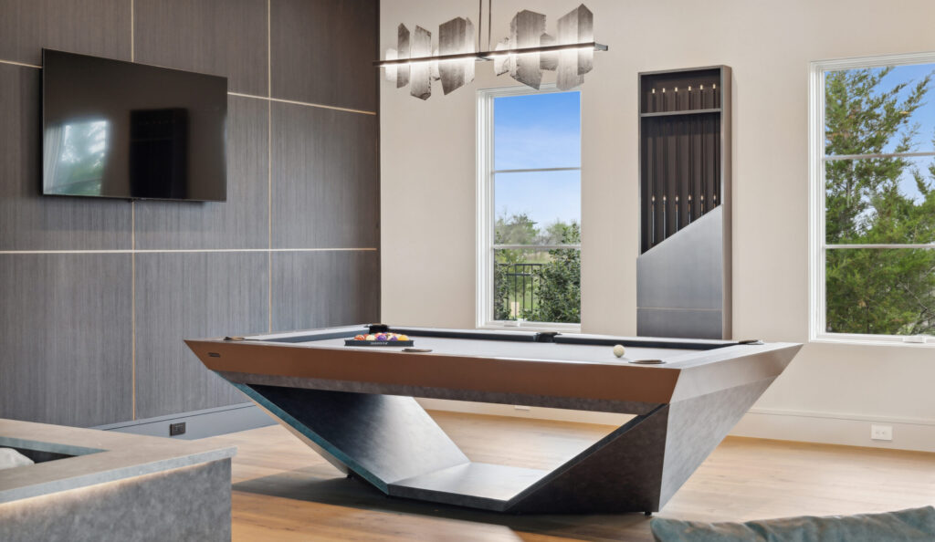 A modern game room exudes elegance with the Stealth Pool Table at its center. It features luxury game tables, a wall-mounted TV, a cue rack, and large windows with tree views, while a contemporary chandelier hangs above the lavish setup.