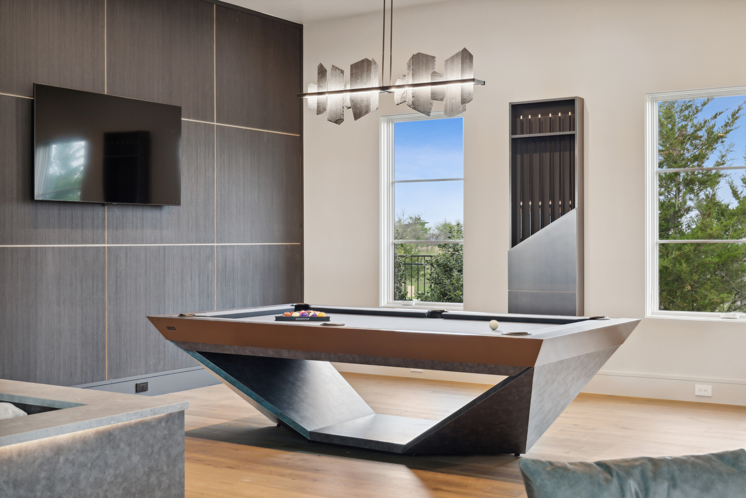 A modern game room exudes elegance with the Stealth Pool Table at its center. It features luxury game tables, a wall-mounted TV, a cue rack, and large windows with tree views, while a contemporary chandelier hangs above the lavish setup.