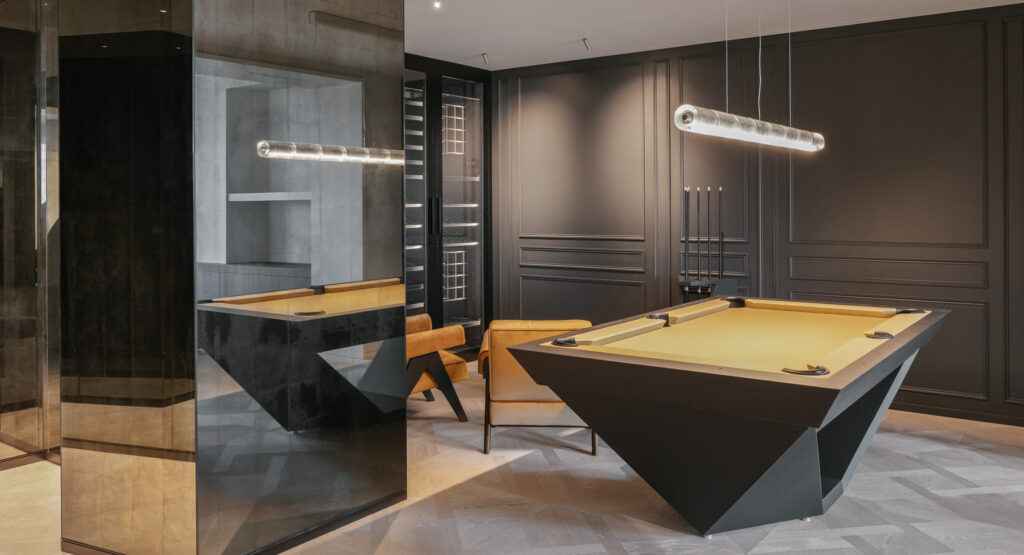 A modern billiard room showcases the luxury Stealth Pool Table in sleek black, paired with stylish brown chairs and a contemporary light fixture. The dark walls feature subtle detailing, creating a sophisticated atmosphere, while a large mirror elegantly reflects part of the space.