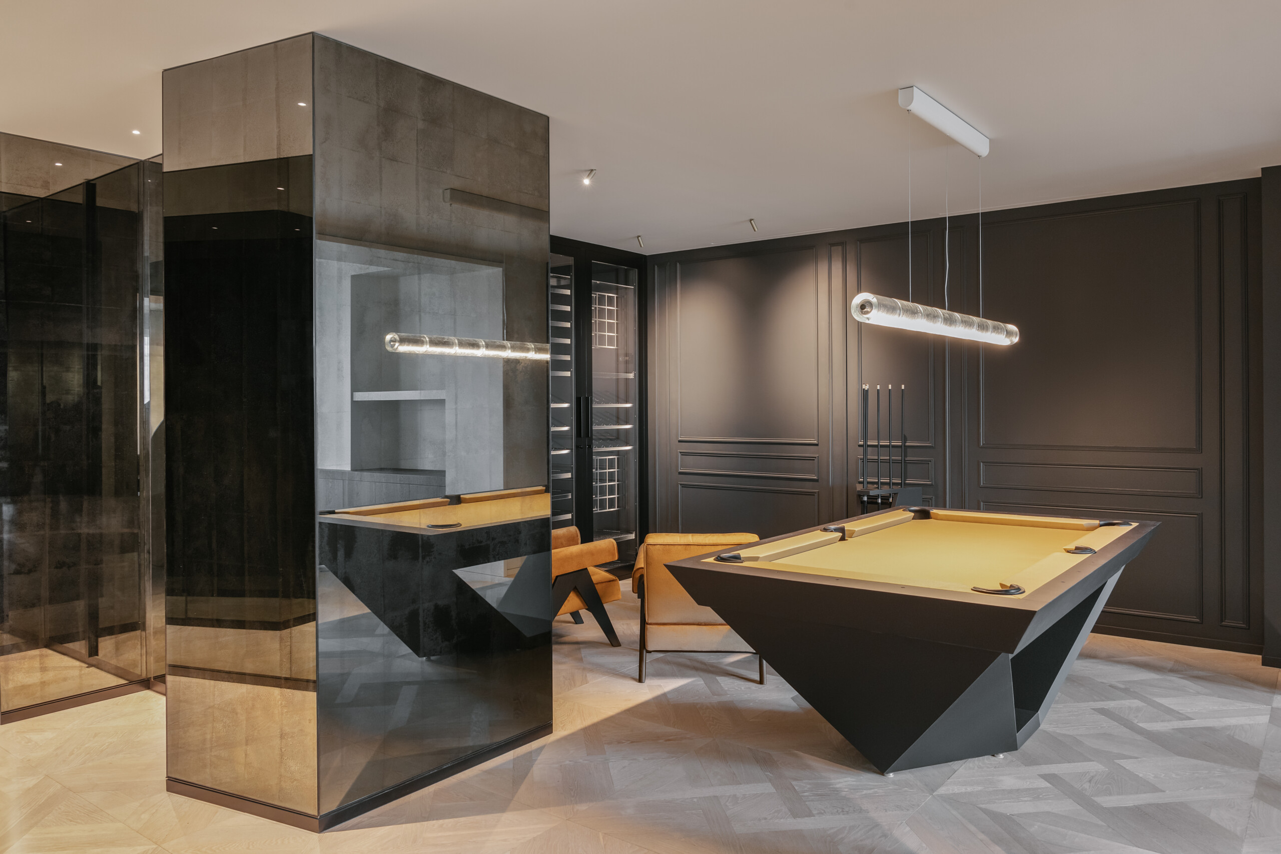 A modern billiard room showcases the luxury Stealth Pool Table in sleek black, paired with stylish brown chairs and a contemporary light fixture. The dark walls feature subtle detailing, creating a sophisticated atmosphere, while a large mirror elegantly reflects part of the space.