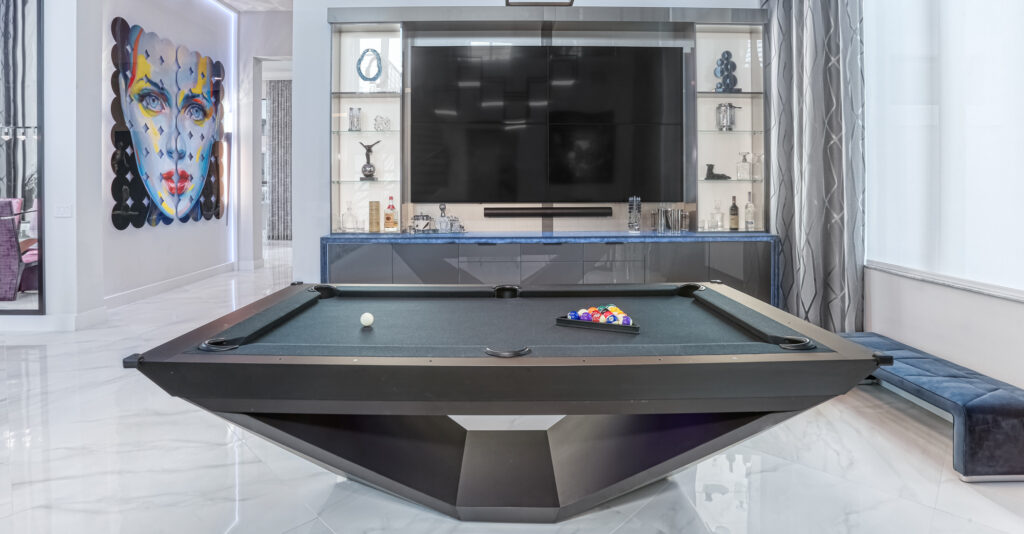A modern game room features the Stealth Pool Table, a luxurious black billiards table with racked balls in front of a large flat-screen TV. The walls display geometric shelving and a colorful abstract portrait, all enhanced by an opulent marble floor.