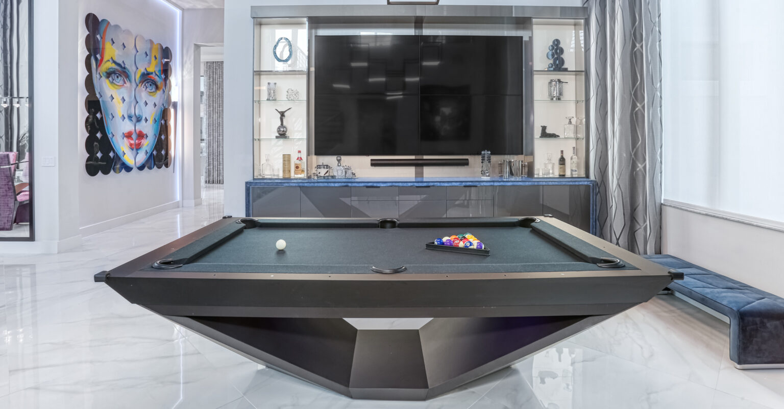 A modern game room features the Stealth Pool Table, a luxurious black billiards table with racked balls in front of a large flat-screen TV. The walls display geometric shelving and a colorful abstract portrait, all enhanced by an opulent marble floor.