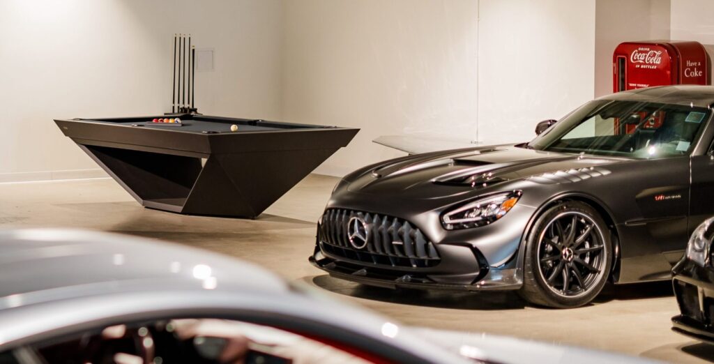 A Stealth Pool Table in sleek black and a gray sports car are parked in a modern, brightly lit showroom, with a vintage red Coca-Cola vending machine visible in the background.