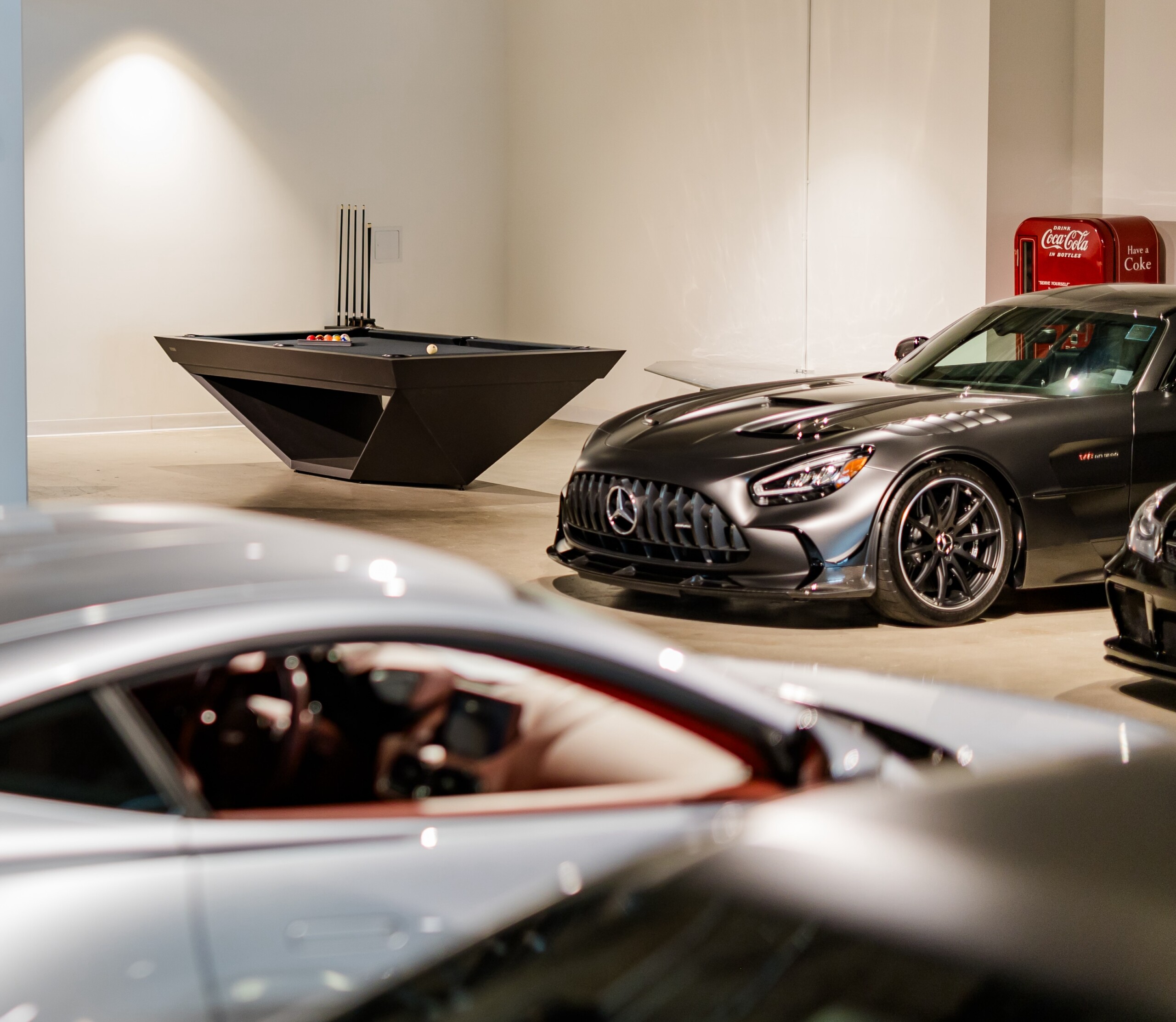 A Stealth Pool Table in sleek black and a gray sports car are parked in a modern, brightly lit showroom, with a vintage red Coca-Cola vending machine visible in the background.