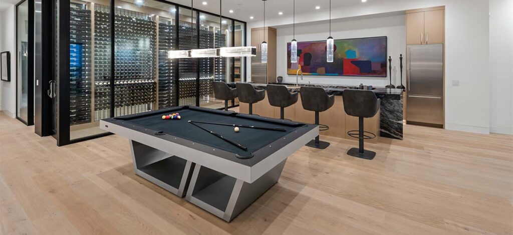 A modern room featuring luxury game tables and a pool table with cues and balls, a sleek bar area with high chairs, and a large wine rack. Contemporary art adorns the walls, while pendant lights illuminate the space.