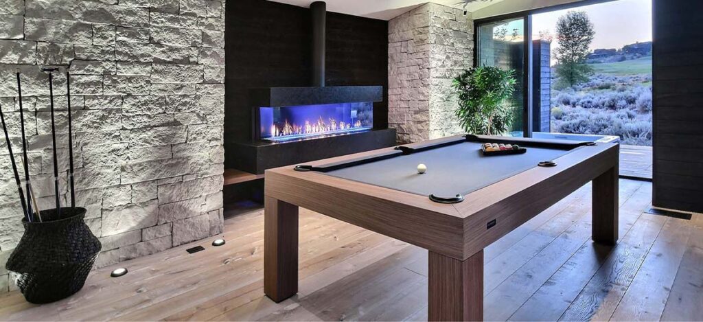 A modern game room features luxury game tables with a pool table at its center, a sleek fireplace in the background, and large glass doors opening to a scenic outdoor view. The room showcases stone walls, wooden flooring, and a potted plant.