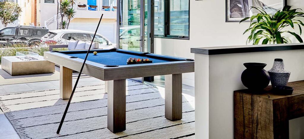A modern room with a luxury game table centered on a striped rug. Pool cues lean against the table, with balls arranged on the blue felt. Large windows offer a view of the patio and street, while decorative plants and pottery adorn the chic space.