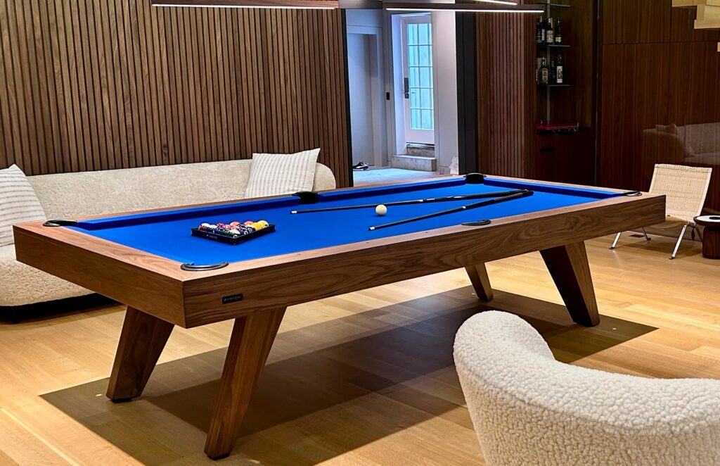 A modern game room featuring a luxury wooden pool table with a blue felt surface. The table is set with a full rack of balls and cues. Surrounding the table are a cream sofa, a white armchair, and wood-paneled walls with shelves.