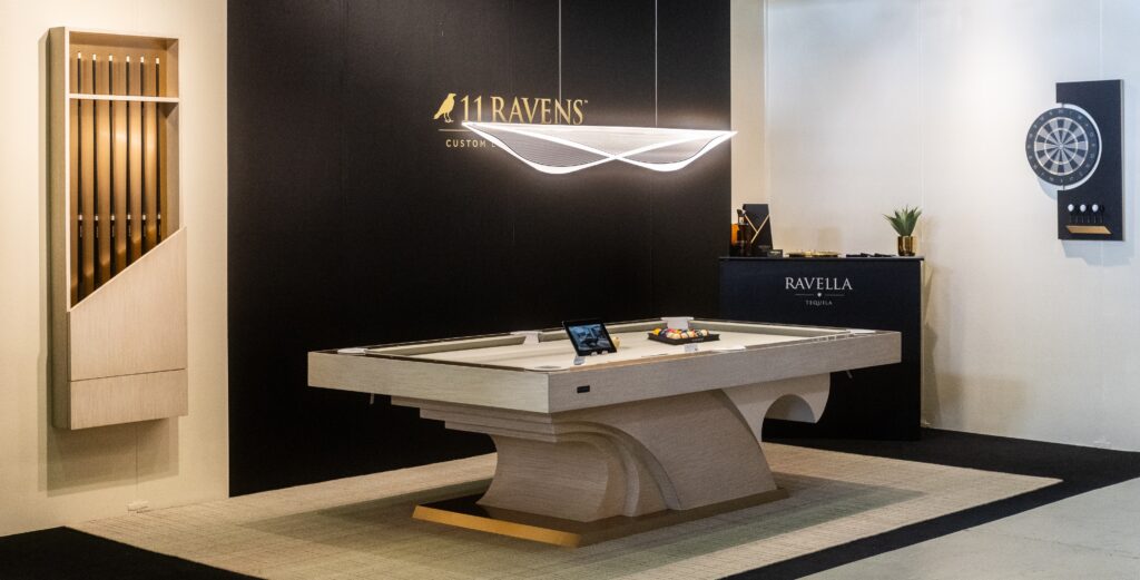The modern showroom showcases the sleek 8 Poseidon Pool Table - Off The Floor with a curved base. Luxurious game tables and a contemporary hanging light fixture set the scene, alongside a dartboard on the wall. A corner counter displays Ravella, contrasting elegantly with light and dark walls.