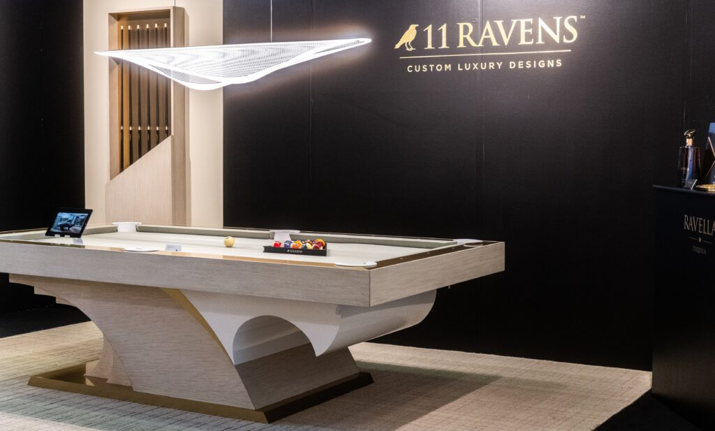 Showcased in a showroom, the 8 Poseidon Pool Table - Off The Floor features a sleek modern curved base with neutral tones, epitomizing 11 Ravens luxury. An elegant geometric light fixture highlights the brands tagline: Custom Luxury Designs.
