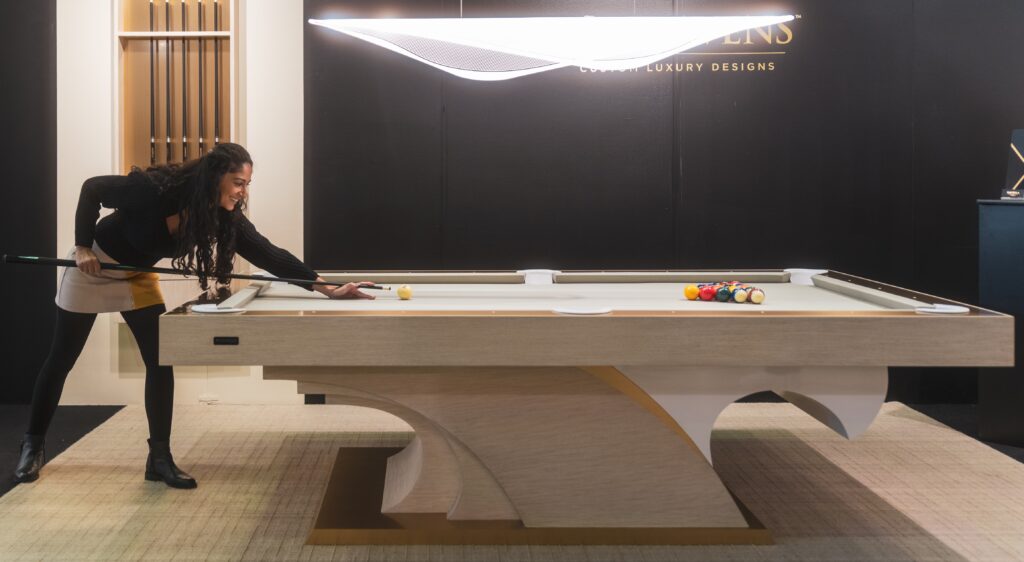 In a stylish interior with soft lighting, a woman with long curly hair aims her cue at the ball on the modern and elegantly designed 8 Poseidon Pool Table - Off The Floor.