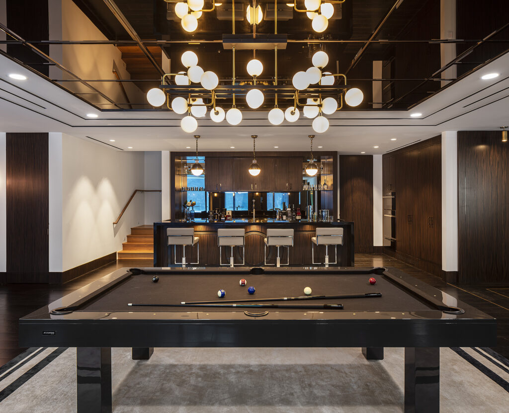 Elegant room featuring a 7 Malibu Pool Table - Off The Floor at its center on a rug, surrounded by sleek wood paneling. A bar with three stools in the background is flanked by shelves filled with various bottles. Overhead, a contemporary chandelier with globe lights illuminates the space.
