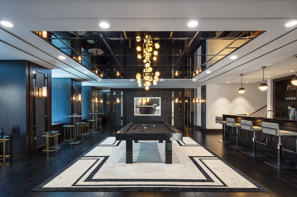 Luxurious game room with a central 7 Malibu Pool Table - Off The Floor on a patterned rug. A modern bar is on the right with stools, while seating areas are to the left. Decorative ceiling lights and mirror accents add elegance to the space.