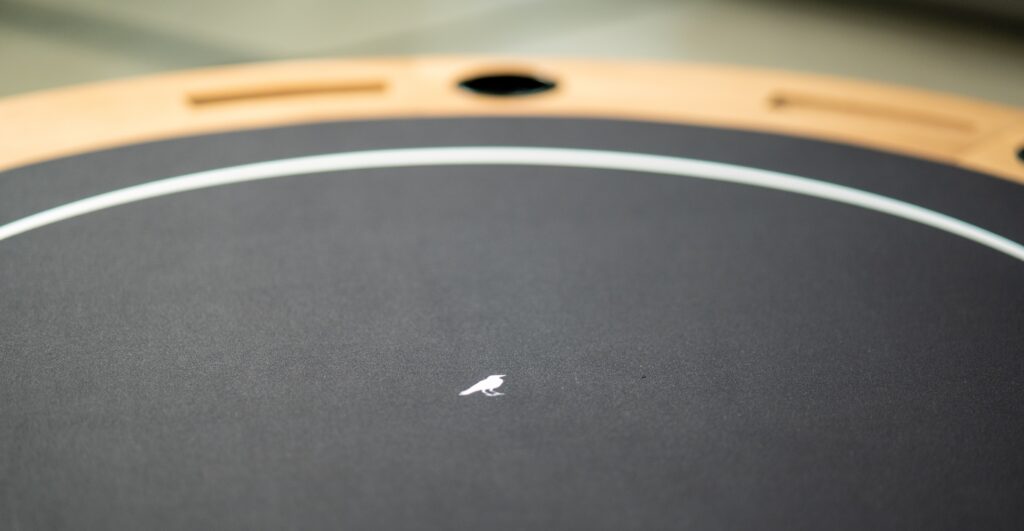 Close-up of the luxurious 5 Theseus Round Poker Table - Off The Floor in black with a light wood edge. It features a small white speck in the center and rounded compartments along its border, seamlessly blending style and function.