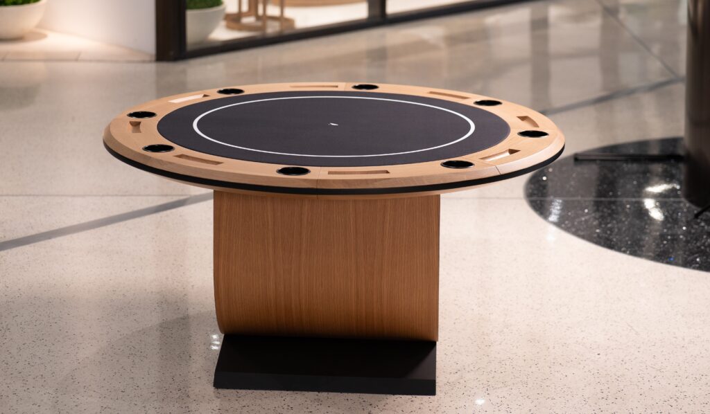 The 5 Theseus Round Poker Table - Off The Floor features a wooden base and sleek black playing surface with multiple cup holders, elegantly placed on a polished indoor floor against a blurred backdrop.