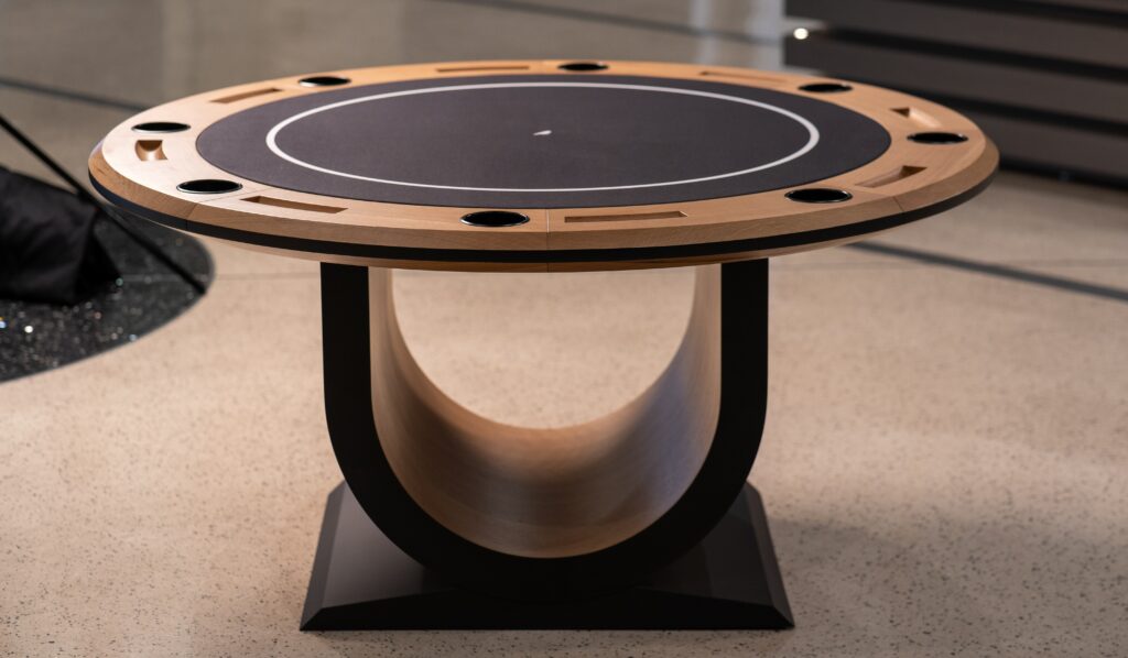 The 5 Theseus Round Poker Table - Off The Floor is a luxurious modern design featuring a black playing surface, wooden outer rim with cup holders, and a distinct curved wooden base, beautifully set on a speckled floor.