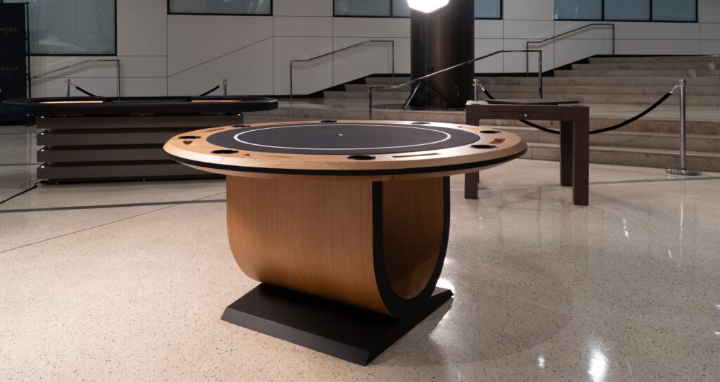 The 5 Theseus Round Poker Table - Off The Floor, with its wooden base and black playing surface, anchors the modern, spacious room as a symbol of luxury. In the background are a staircase, rope barrier, and smaller furniture piece.