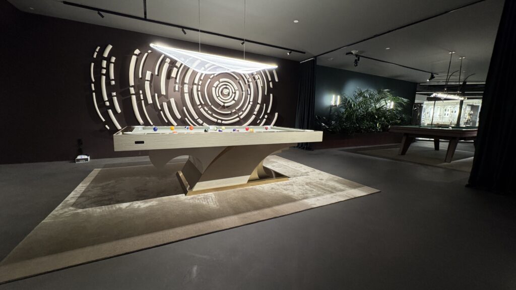 A modern game room features the 8 Poseidon Pool Table - Off The Floor, illuminated by a sleek overhead light. An abstract circular design adorns the wall, and plants in the corner add greenery to the stylish, dark-toned space.