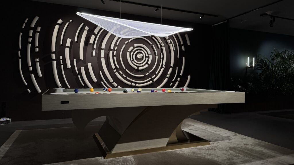 In a dimly lit room, the 8 Poseidon Pool Table - Off The Floor stands elegantly with billiard balls artfully arranged on its surface. A circular, abstract white design graces the wall, while a unique sculptural light fixture casts an elegant glow over the scene.
