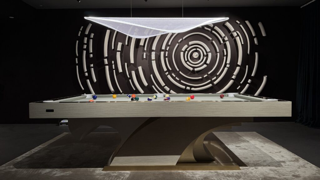 The 8 Poseidon Pool Table - Off The Floor, featuring vibrant billiard balls, stands on a chic rug. Overhead, a modern light fixture adds a sophisticated glow. The wall displays a circular abstract art piece in sleek black and white tones.