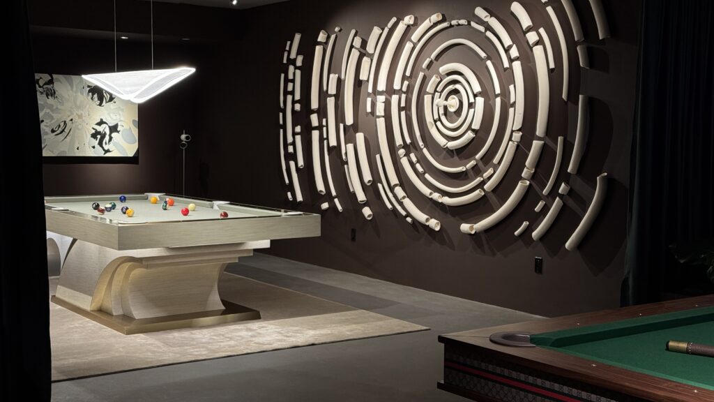 A chic indoor space highlights the 8 Poseidon Pool Table - Off The Floor, complete with vibrant balls. The ambiance is enriched by dim lighting, a striking abstract circular wall sculpture, and a unique geometric light fixture casting soft illumination.