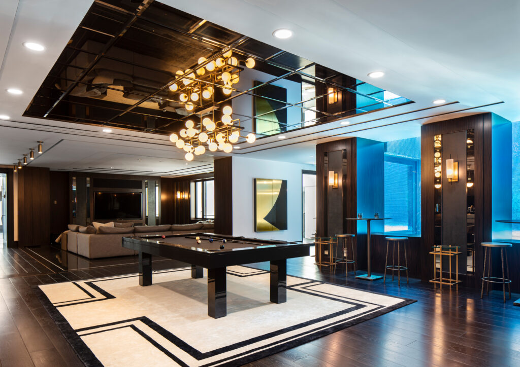 A luxurious game room features the sleek 7 Malibu Pool Table - Off The Floor on a stylish carpet. A large chandelier reflects above, enhancing the lounge area with its sofa, decorative lighting, and blue accent panels on the walls.