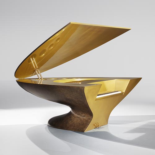 A modern, sculptural grand piano with a gold and brown finish, featuring a sleek, curved base and an open, angular lid. The piano has a futuristic design and stands on a plain, light background.