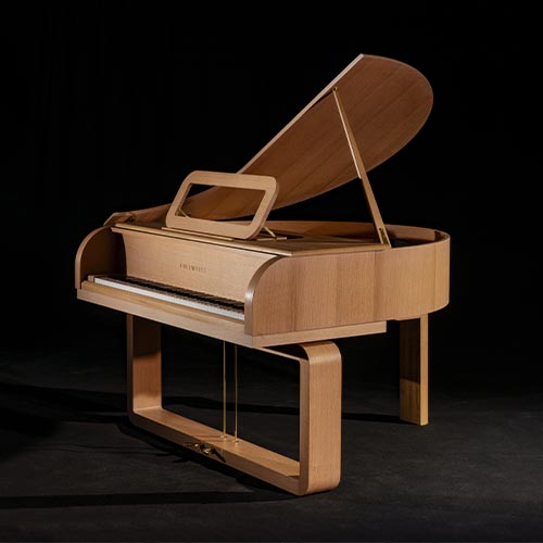 A modern grand piano made of light wood with a unique, minimalist design and rounded edges, set against a black background. The lid is open, and the piano has a single supported leg and a rectangular base.