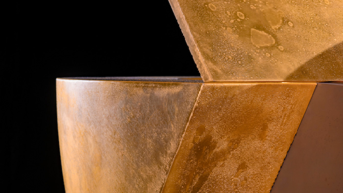 A close-up of two angular, metallic gold sculptures with textured surfaces, stacked against a black background—evoking the bold elegance of Edelweiss Pianos.