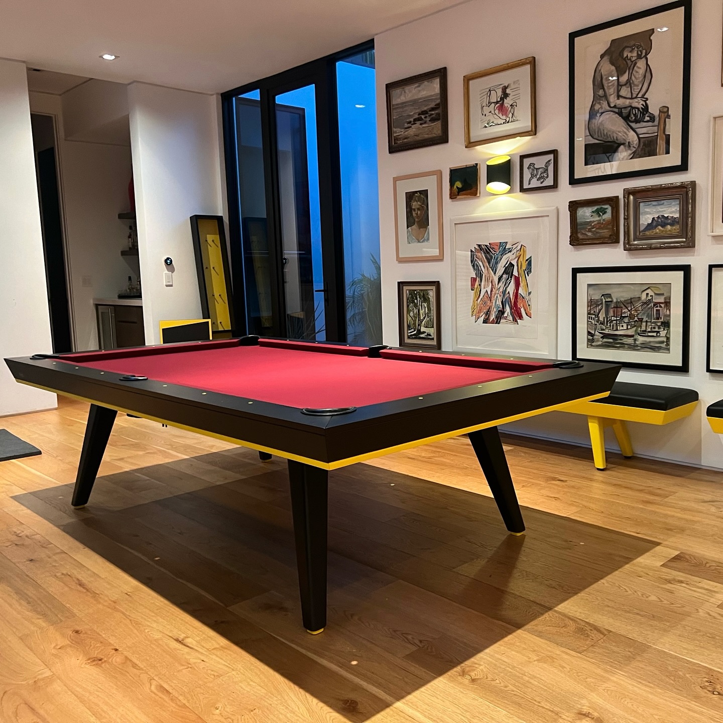 A red-and-black Hamptons Pool Table with Simonis cloth and yellow accents stands in a modern room with wooden floors, large windows, and walls adorned with framed artworks reminiscent of Beverly Hills style.