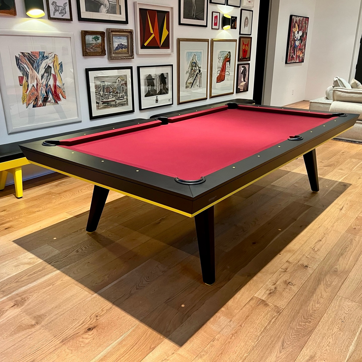 A modern Hamptons Pool Table with vibrant red Simonis cloth and a black-and-yellow frame sits on a wooden floor, complemented by a gallery wall of colorful framed art, evoking Beverly Hills style.