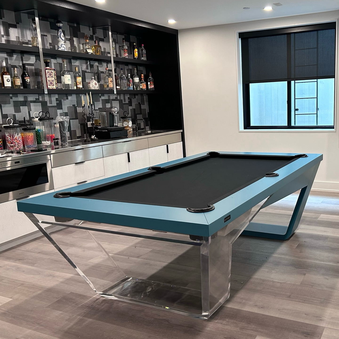 A modern room with a sleek blue and clear acrylic pool table, wooden floors, a wall of cabinets, shelves stocked with liquor bottles, and a window with a black roller shade.