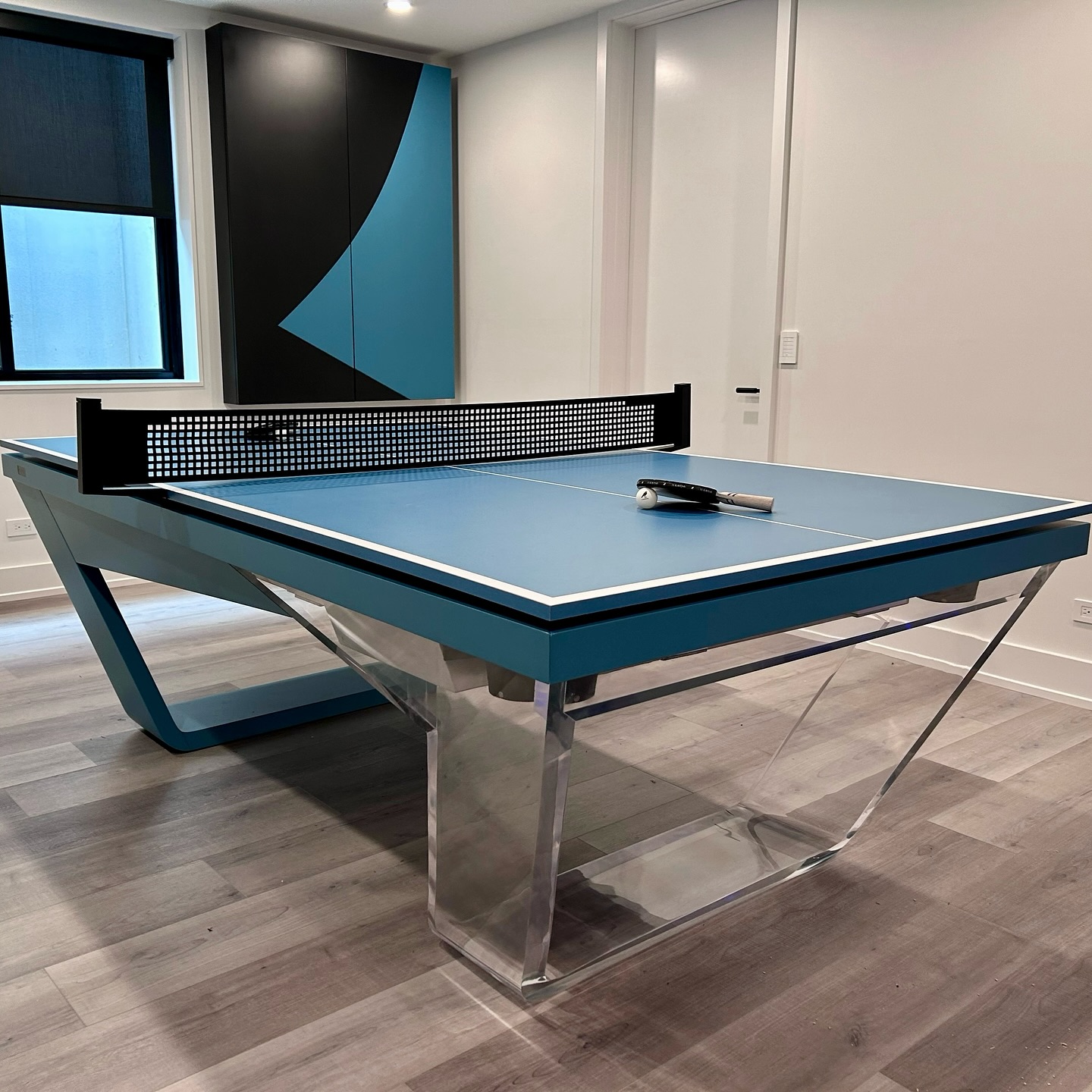 A modern blue ping pong table with a transparent acrylic base and a black net sits in a stylish room with light wood floors and geometric wall decor. Two paddles and a ball are on the table.