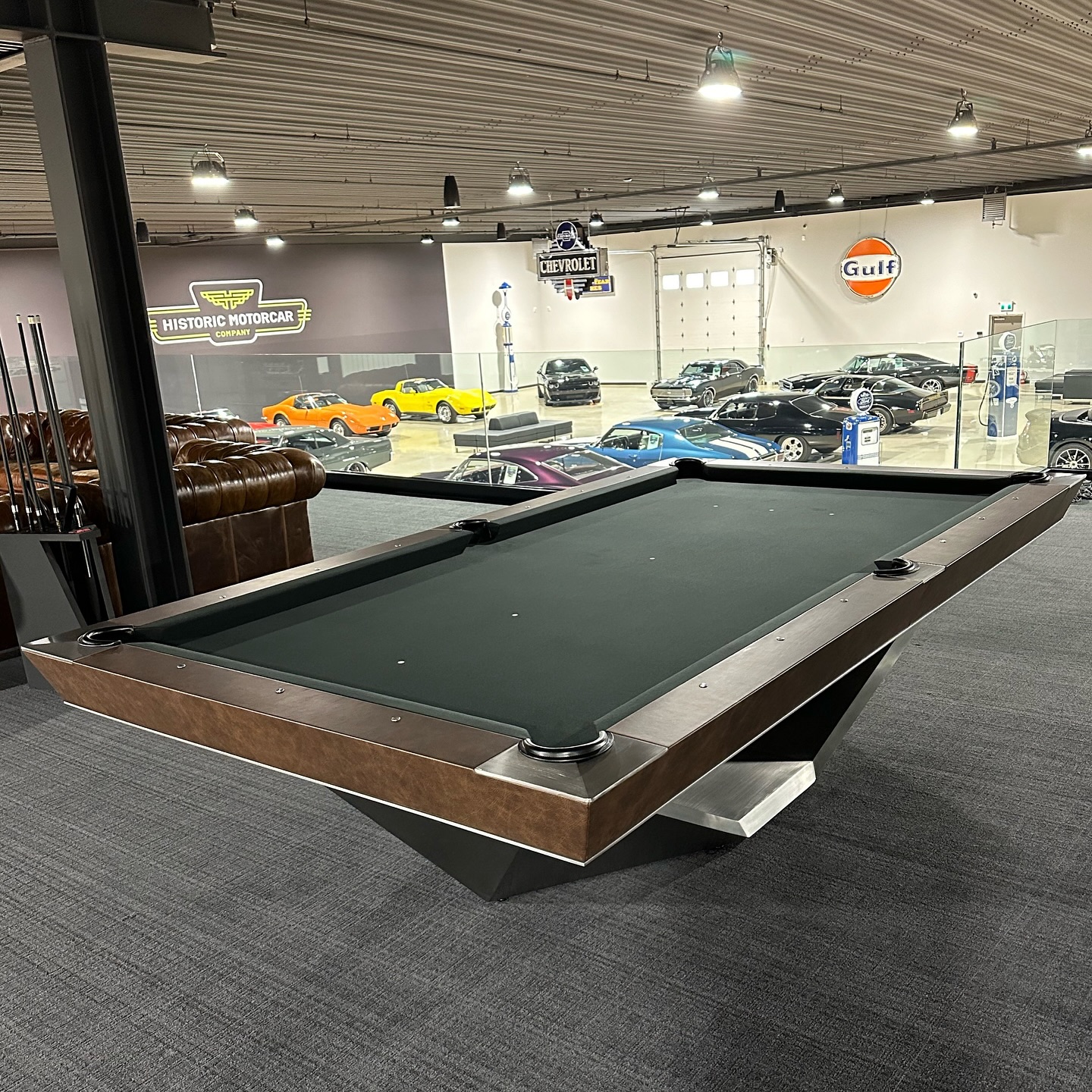 A modern pool table sits on a carpeted floor overlooking a showroom filled with classic cars, including yellow, orange, blue, and black vehicles, with vintage signs on the walls.