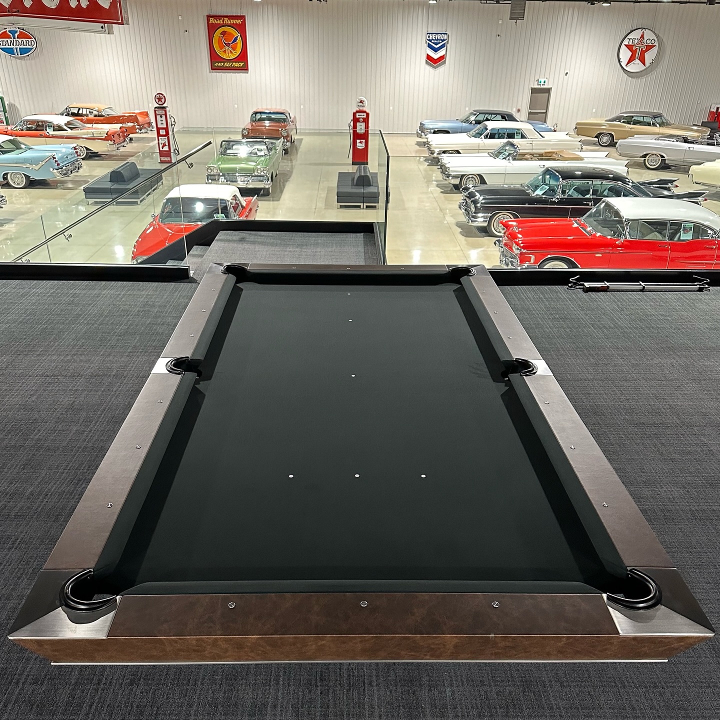 A black pool table is in the foreground, with a collection of vintage cars, old gas pumps, and nostalgic signs visible in the background within a spacious indoor showroom.