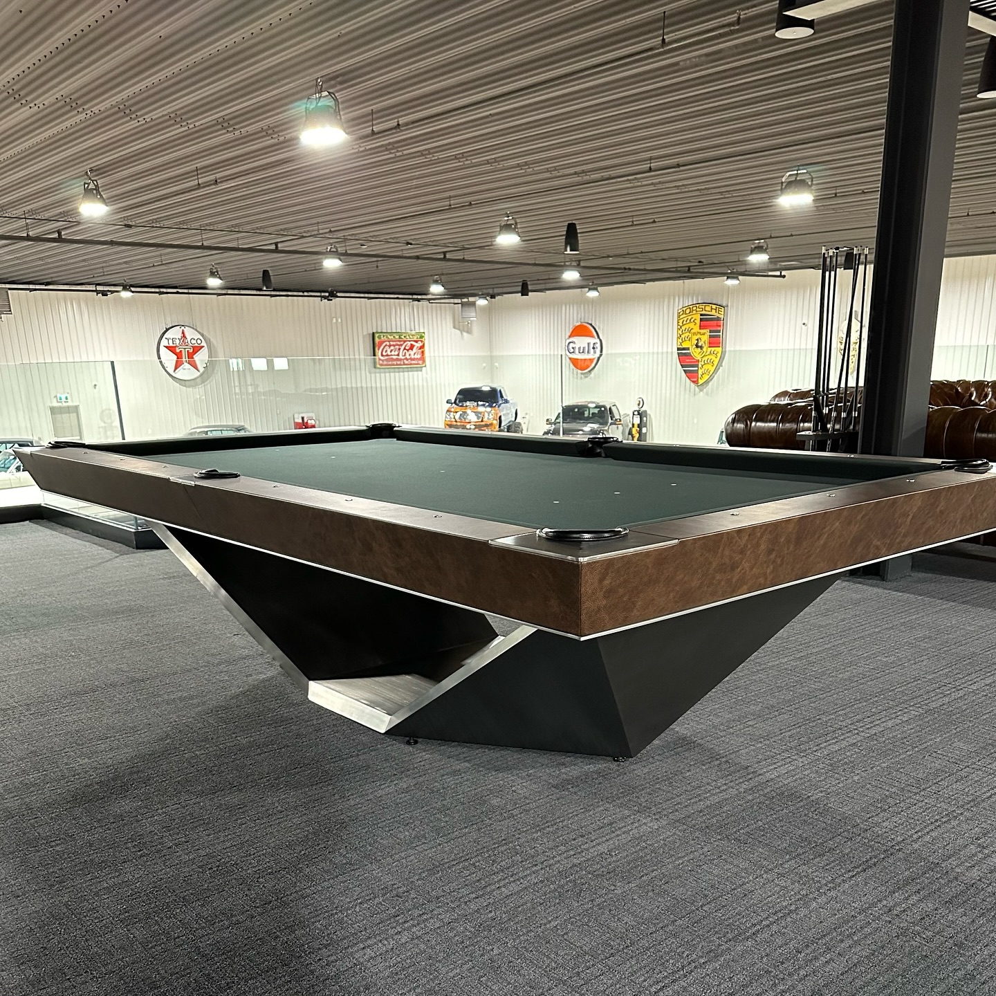 A modern pool table with a geometric base stands in a spacious room with gray carpet. Classic car-related signs and several cars are visible in the background under bright ceiling lights.