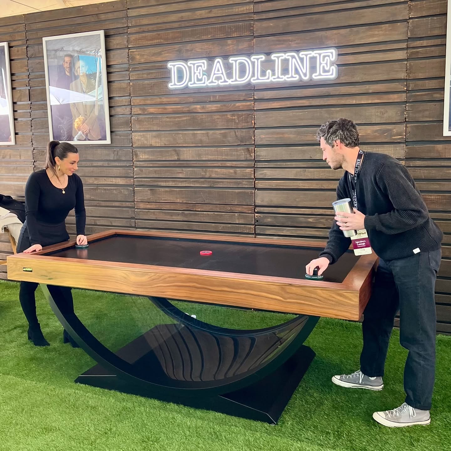 Two people play shuffleboard on a modern wooden table with a curved black base. A neon DEADLINE sign glows on a wooden wall behind them. The floor is covered in green turf.