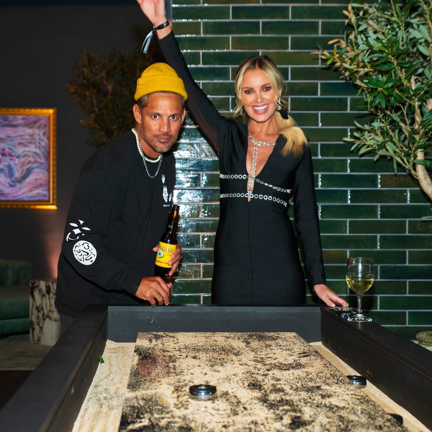 A man in a yellow beanie and a woman in a black dress stand beside a shuffleboard table, smiling. The man holds a drink, while the woman raises her arm in celebration. They are indoors, in front of a green tiled wall.
