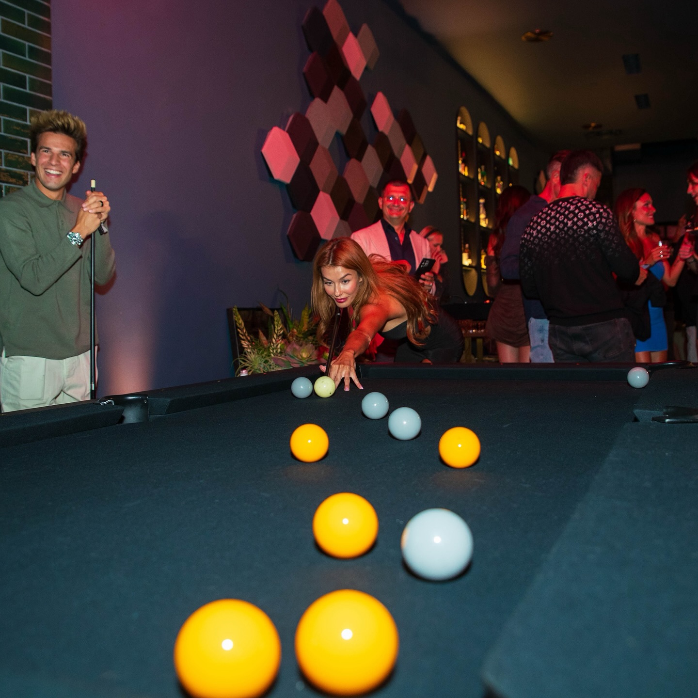 A woman leans over a black pool table to take a shot, surrounded by yellow and white balls, while people watch and socialize in a colorful, lively bar or club setting.