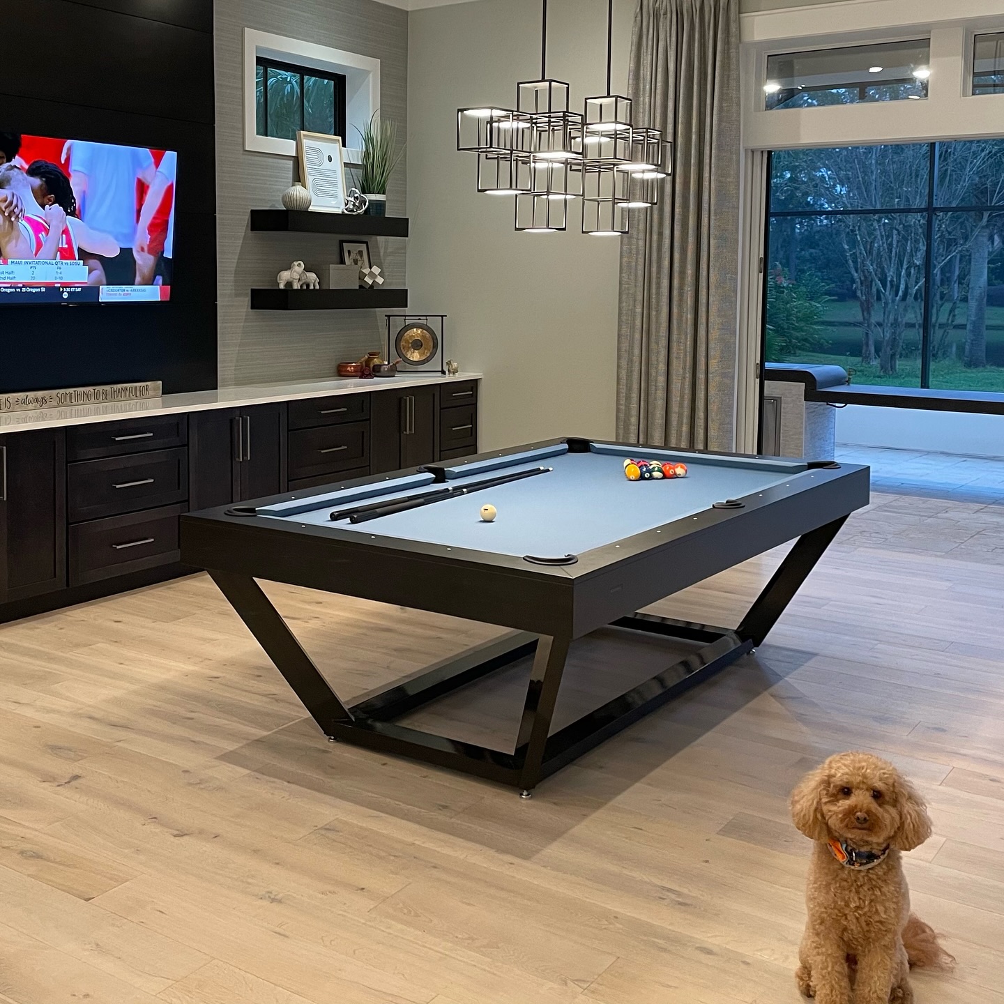 A modern living room with a pool table, balls set for a game, a TV showing sports, dark cabinets, decorative shelves, large windows, and a small brown dog sitting on the wooden floor.