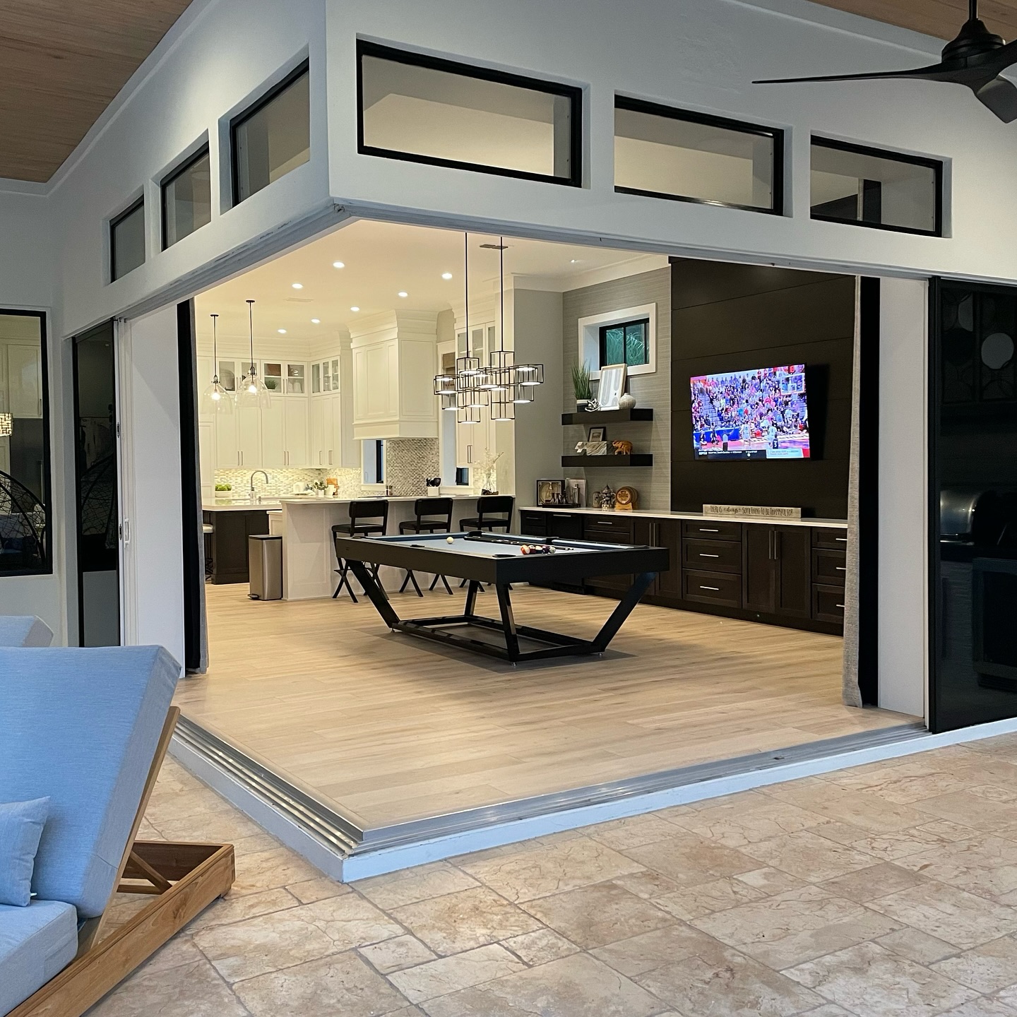 Modern open-concept kitchen and dining area with sliding glass doors, a long dining table, contemporary lighting, wall-mounted TV, and views of an outdoor patio with tiled flooring and lounge furniture.