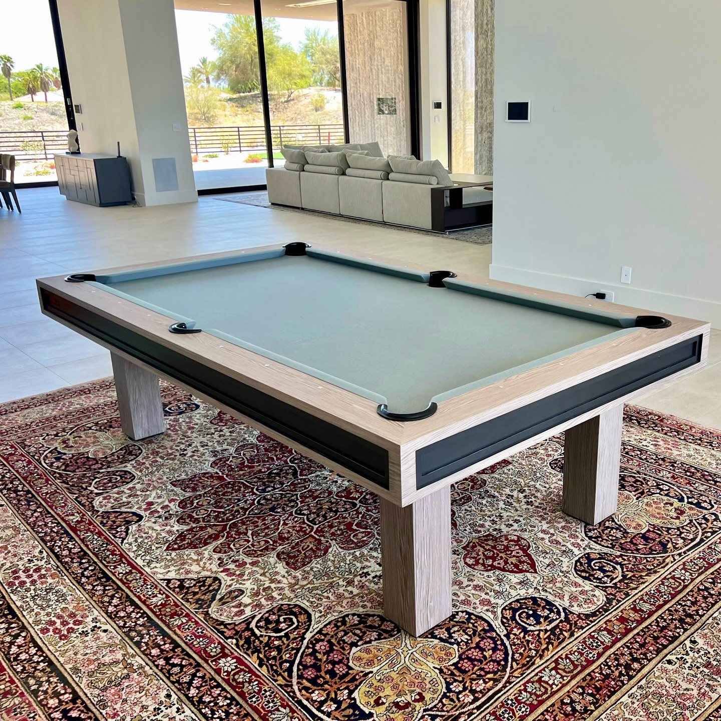 A modern pool table with a light wood frame and gray felt stands on a patterned rug in a spacious, bright room with large windows and contemporary furnishings.