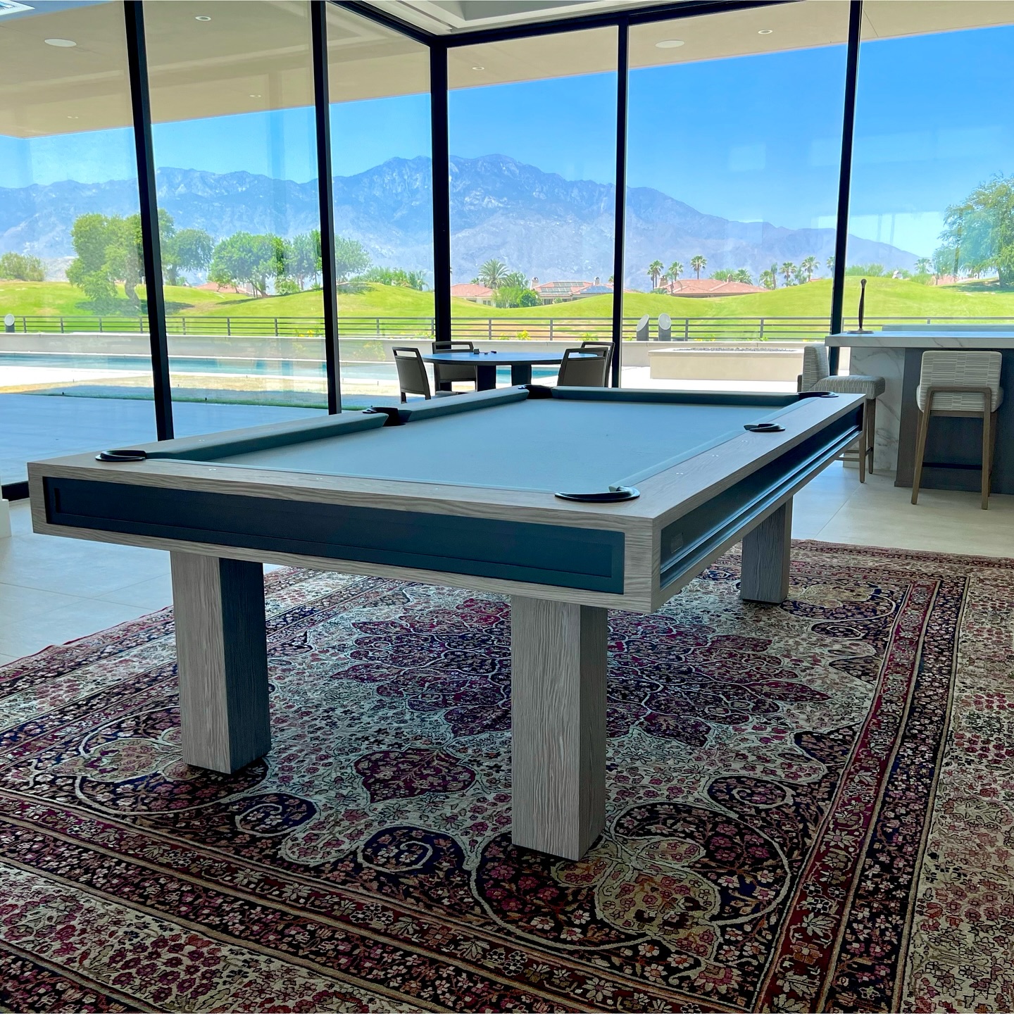 A modern billiards table sits on a patterned rug in a bright room with floor-to-ceiling windows, offering a scenic view of mountains, palm trees, and a golf course outside.