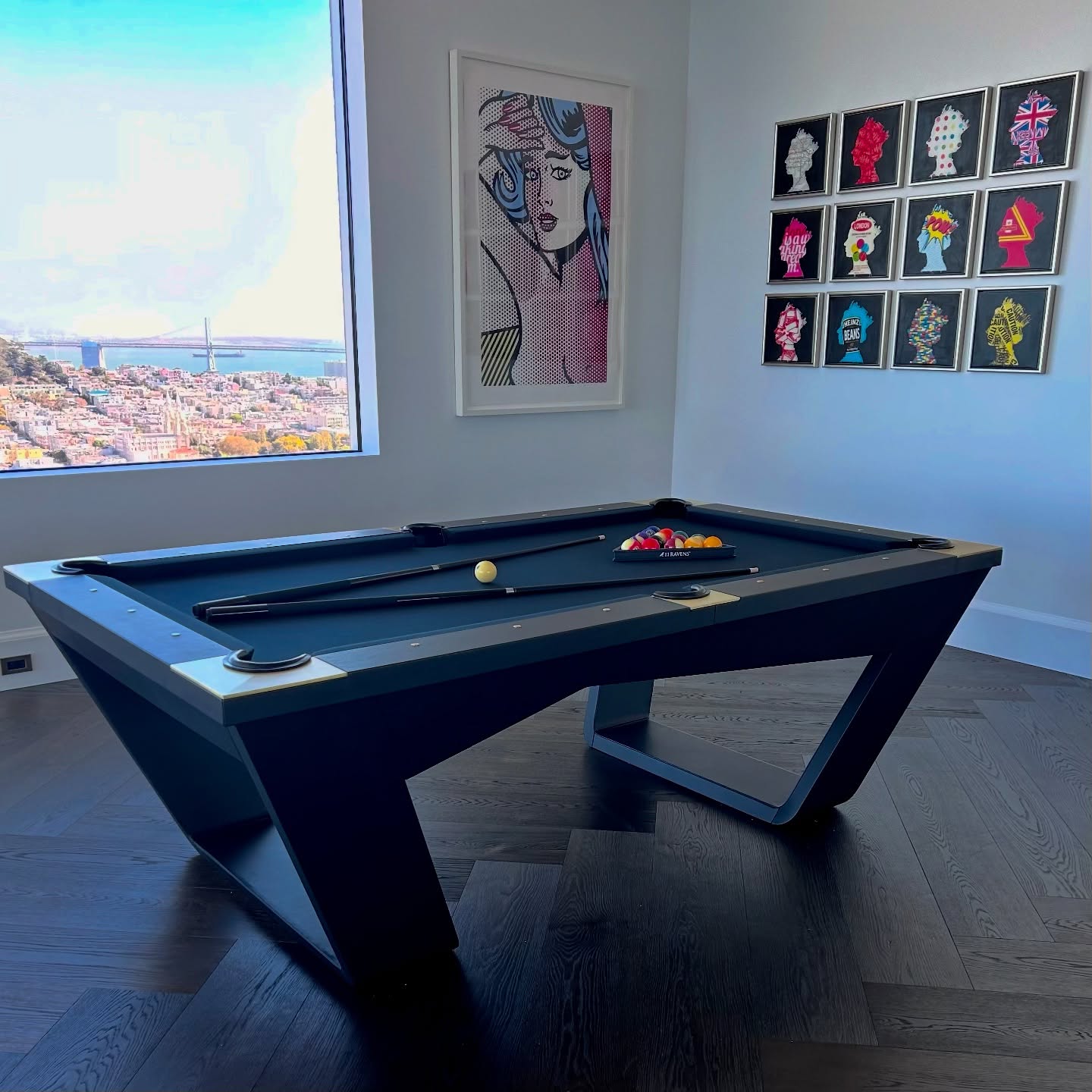 A modern pool table is set in a stylish room with wood flooring, colorful artwork on white walls, and a large window offering a cityscape view with a bridge and water in the distance.