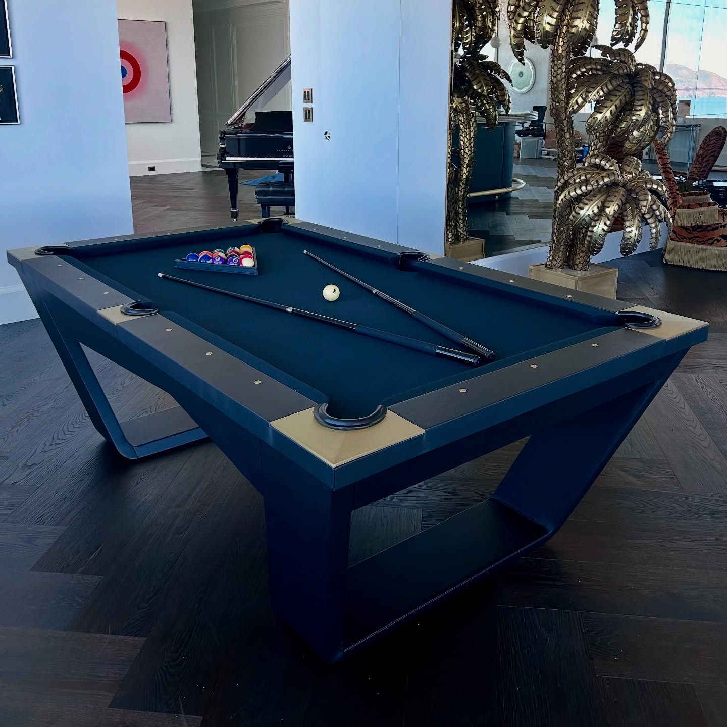 A modern black pool table with balls racked and two cue sticks on its surface stands in a stylish room with dark wood floors, a grand piano, decorative palm sculptures, and contemporary furniture.