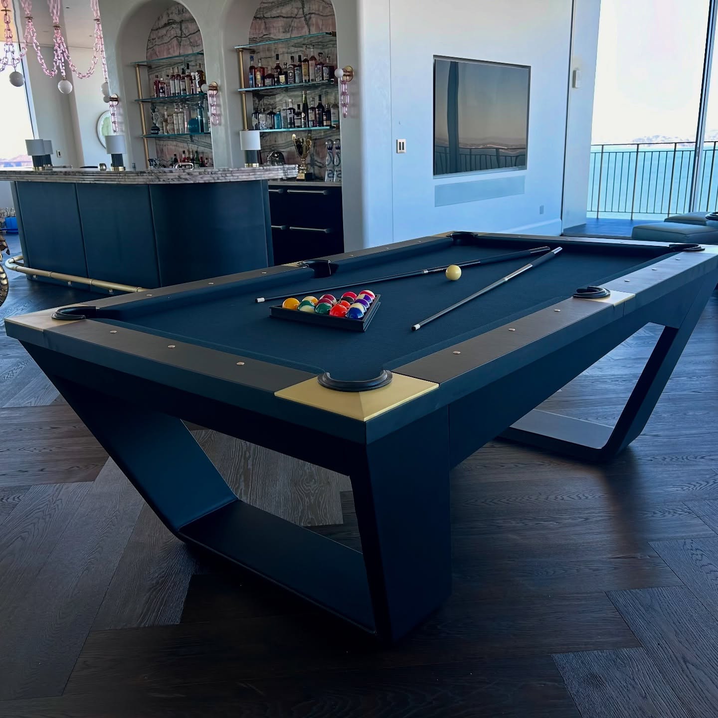 A modern pool table with black felt stands in a stylish room with dark wood floors, a sleek bar in the background, and large windows offering a scenic view. Pool cues and balls are arranged on the table.