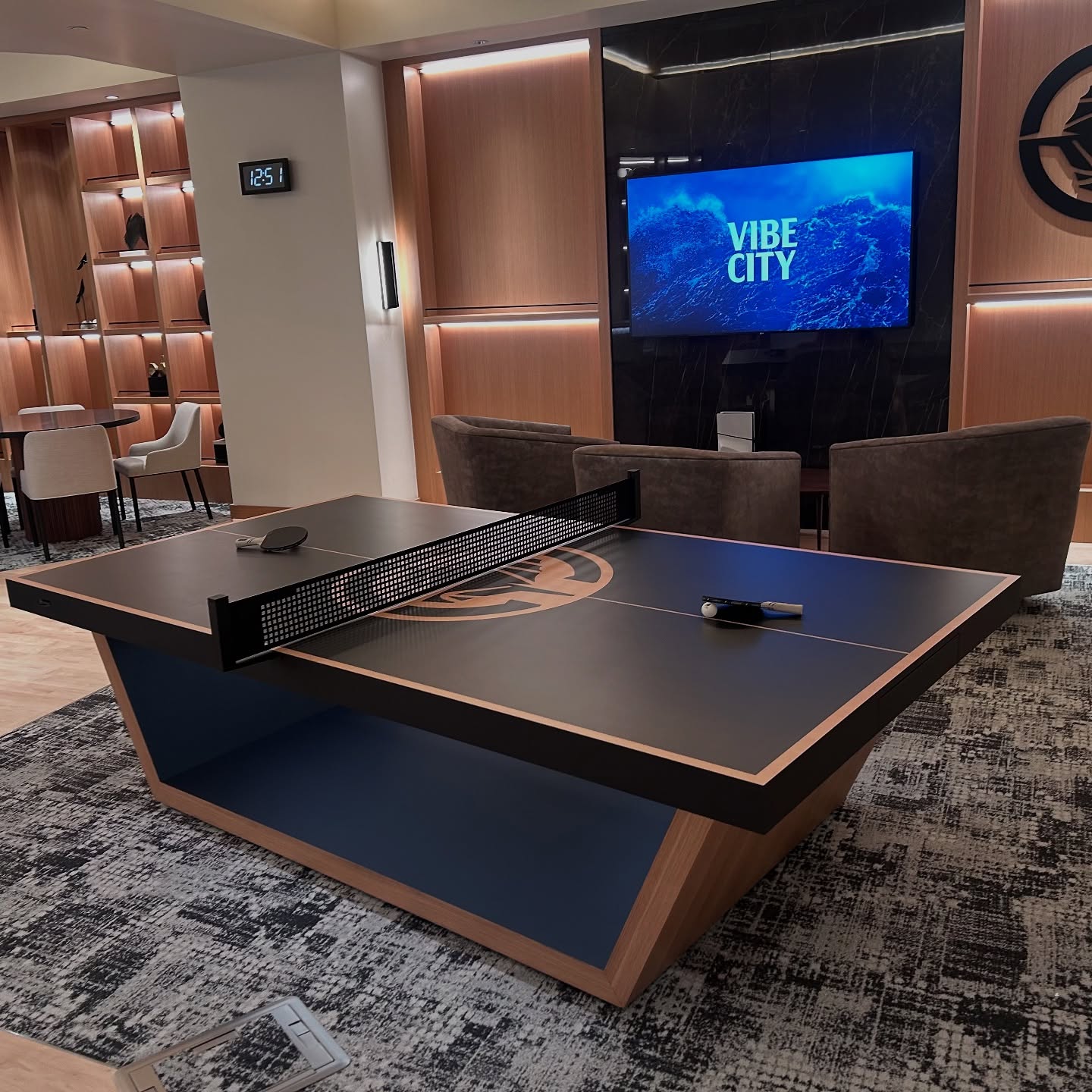 A modern lounge with a stylish ping pong table in the center, two paddles and a ball on the table, chairs and tables in the background, and a TV on the wall displaying VIBE CITY.