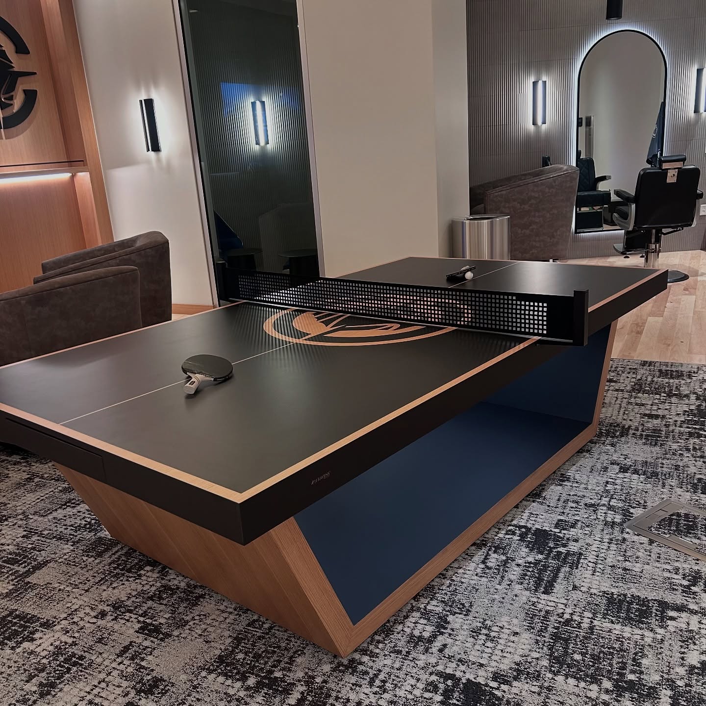 A modern black and wood ping pong table with paddles and a net is set in a stylish lounge with armchairs, wall lights, and a patterned carpet. A large mirror and salon chair are visible in the background.